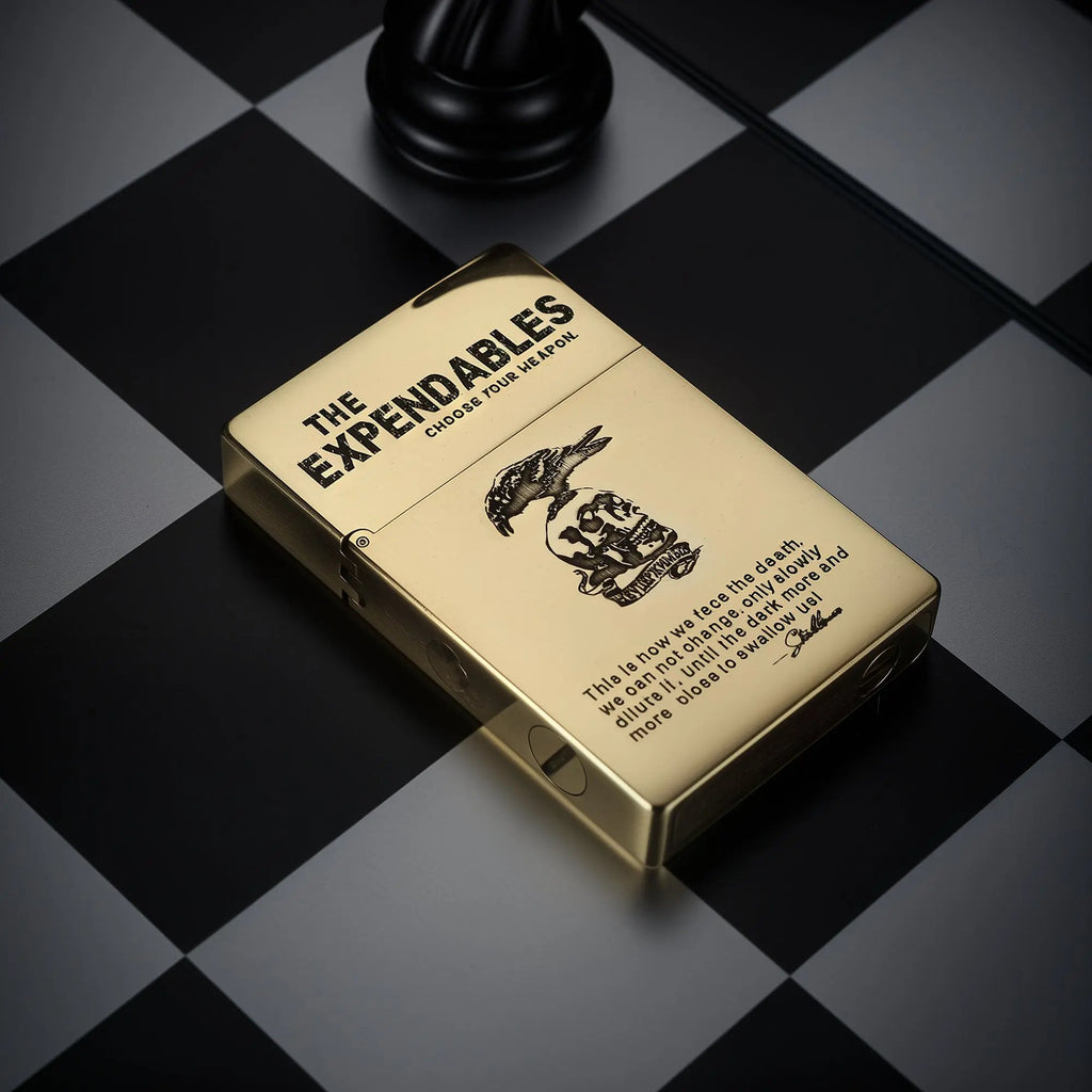 Polished gold-tone Thorens lighter with THE EXPENDABLES etched logo and skull design, displayed on a black and white checkered chessboard.