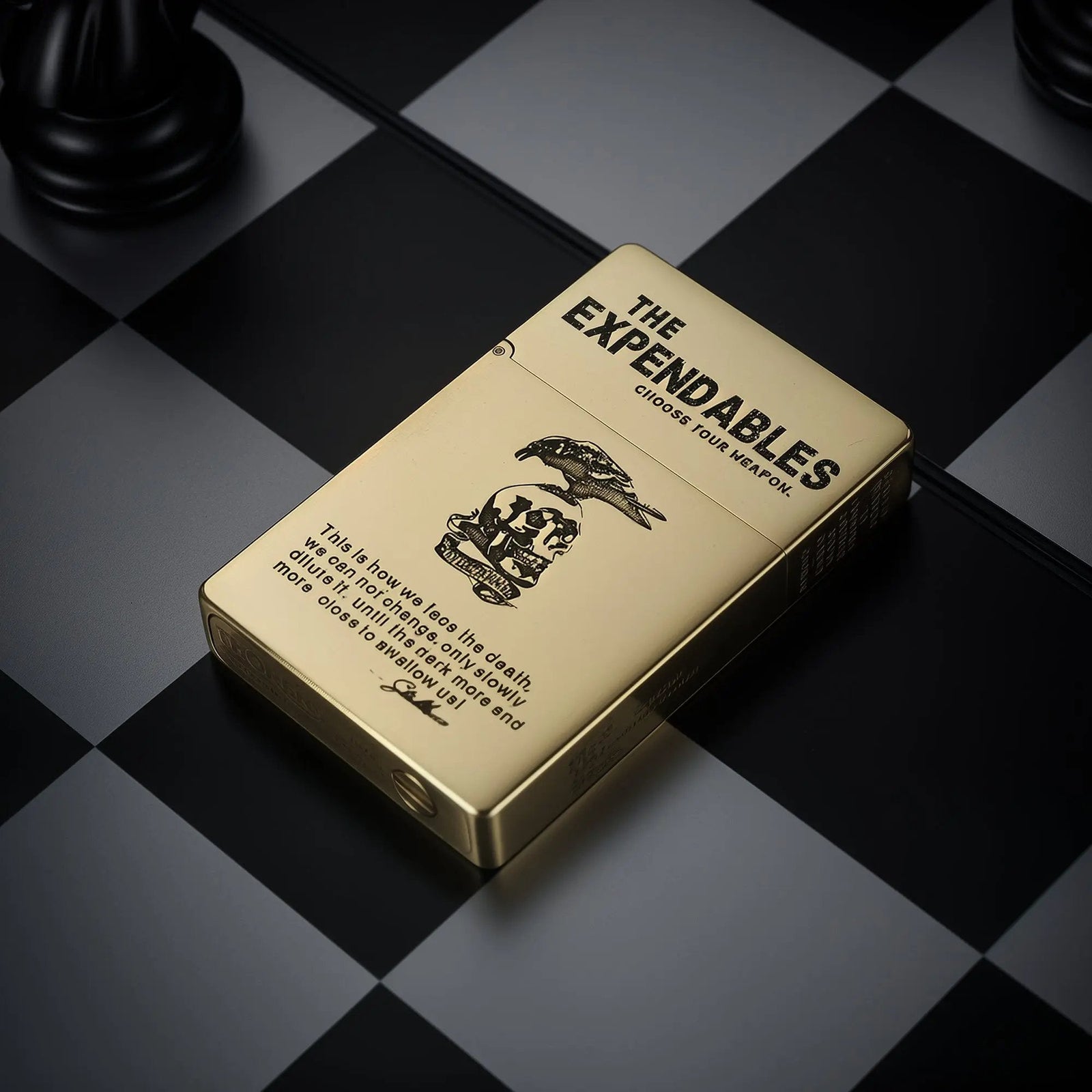 Polished brass Thorens lighter featuring The Expendables logo and skull etching, positioned on a classic black and white checkered surface.