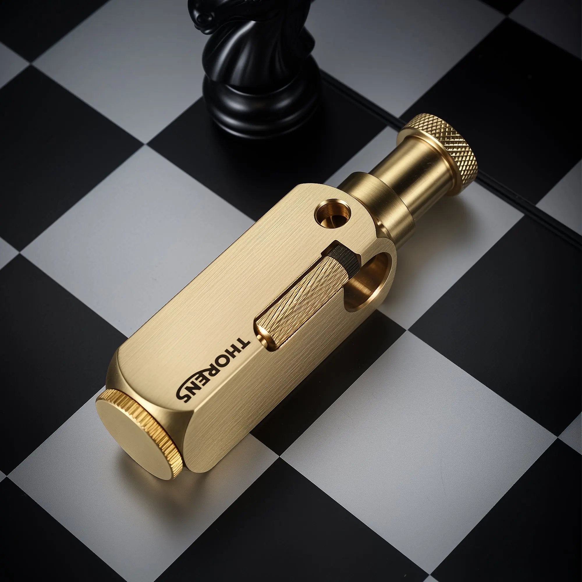 A brushed brass Flamido lighter with a unique octagonal body, featuring the Thorens logo and a knurled plunger, resting on a black and white chessboard.