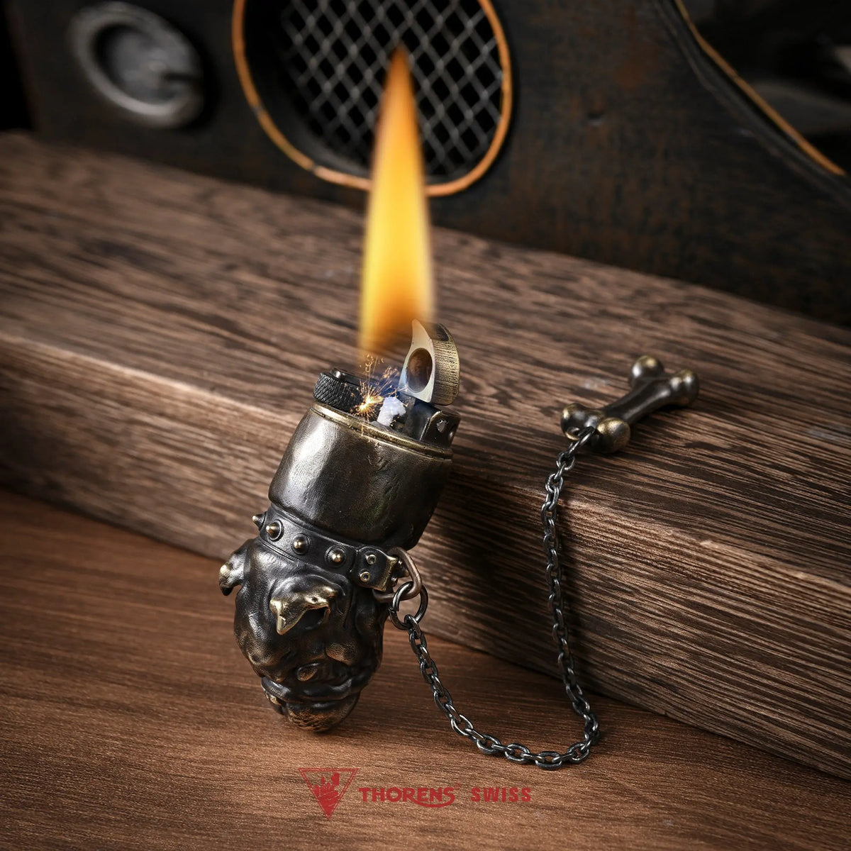 An angled close-up of the Thorens French Bulldog lighter in antique brass with a lit flame, featuring a detailed dog head sculpture, studded collar, and a bone charm on a chain.