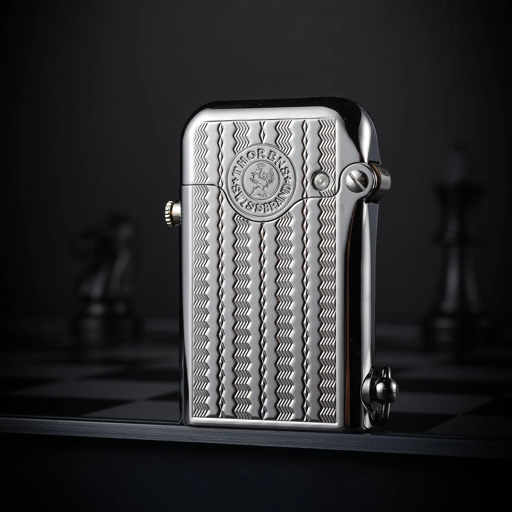 A closed silver Gen 1 Single Claw lighter with an intricate water ripple engraving and a Thorens Swiss emblem, standing on a dark chessboard.