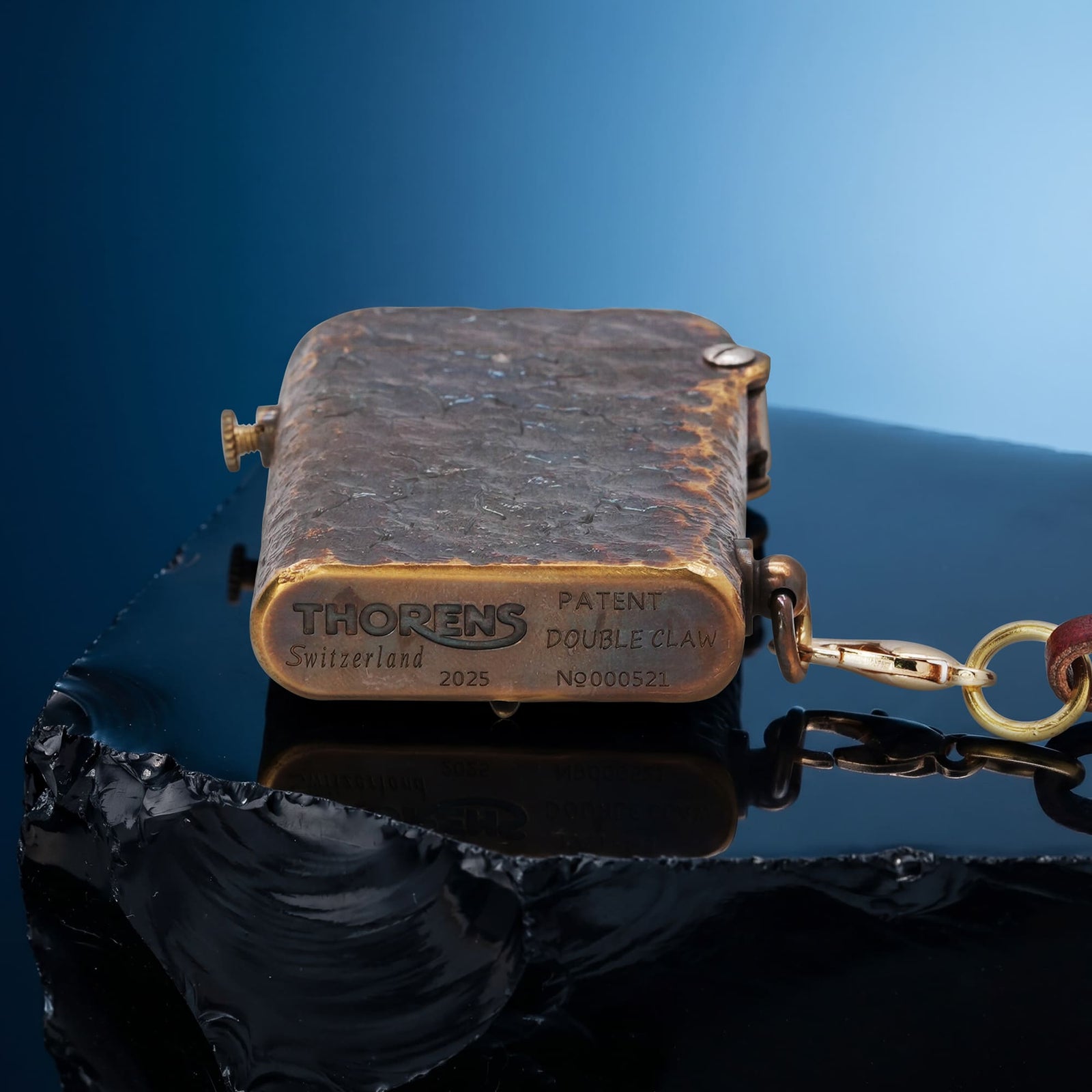 Thorens Gen 2 Double Claw Dark Angel Blade lighter shown closed on a reflective black surface, highlighting engraved base markings, distressed brass texture, and side chain.