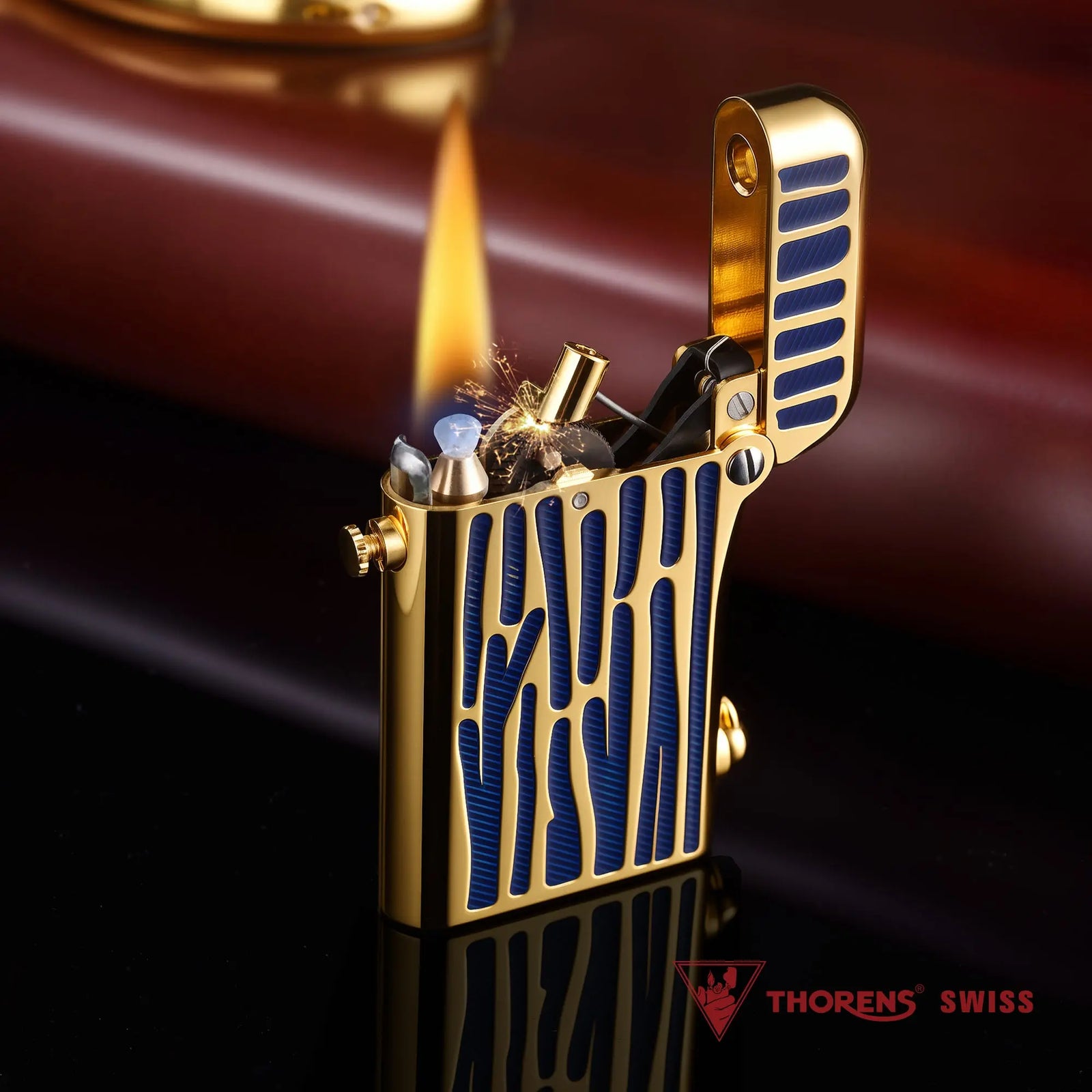 An ignited Thorens Gen 2 Double Claw mechanical lighter with gold finish and blue textured stripes, displaying the internal spring mechanism, flint sparks, and the "Thorens Swiss" logo in the foreground.