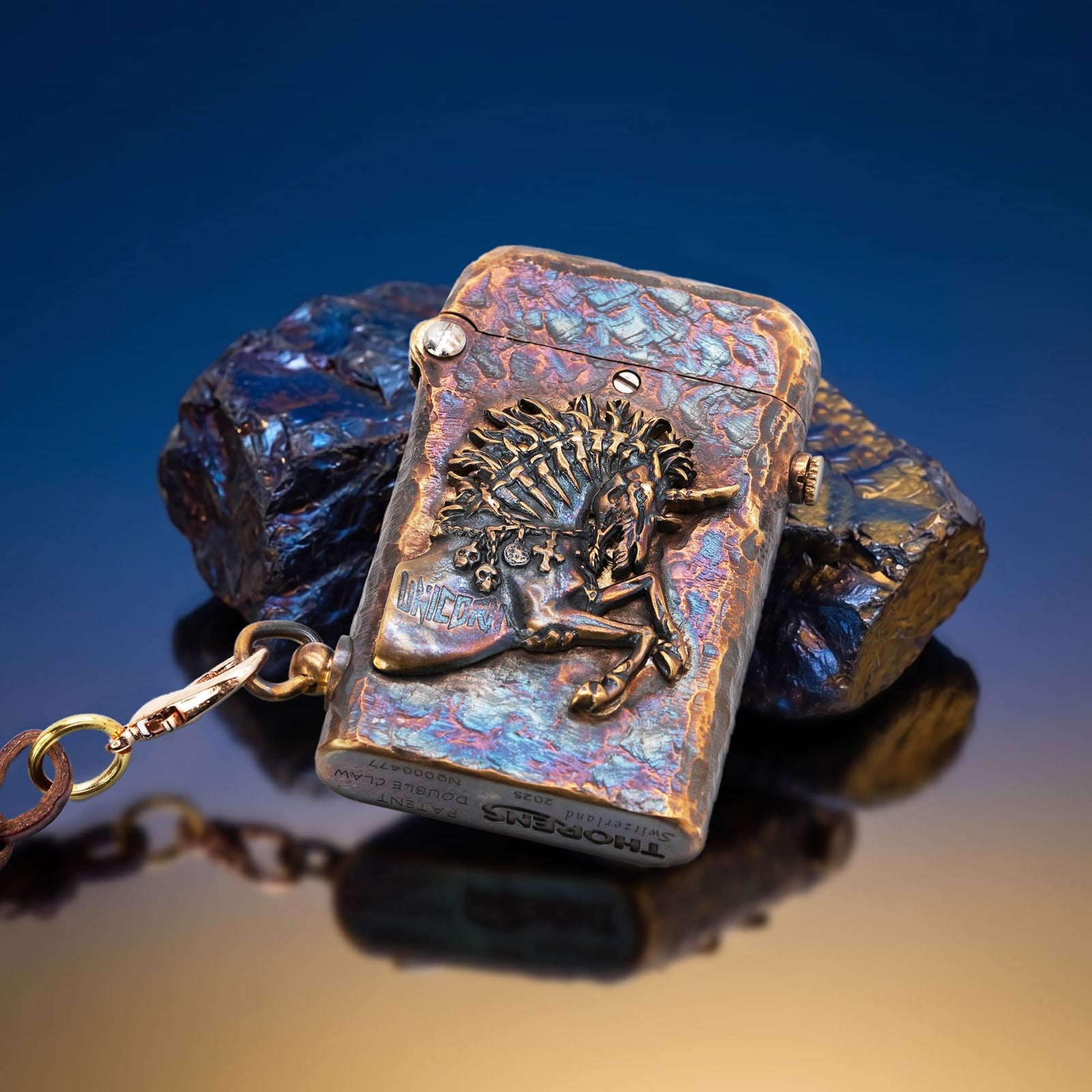 Thorens Gen 2 Double Claw Warhorse lighter displayed closed on a reflective surface beside dark stones, featuring a multitone distressed finish, raised warhorse motif, and attached leather lanyard.
