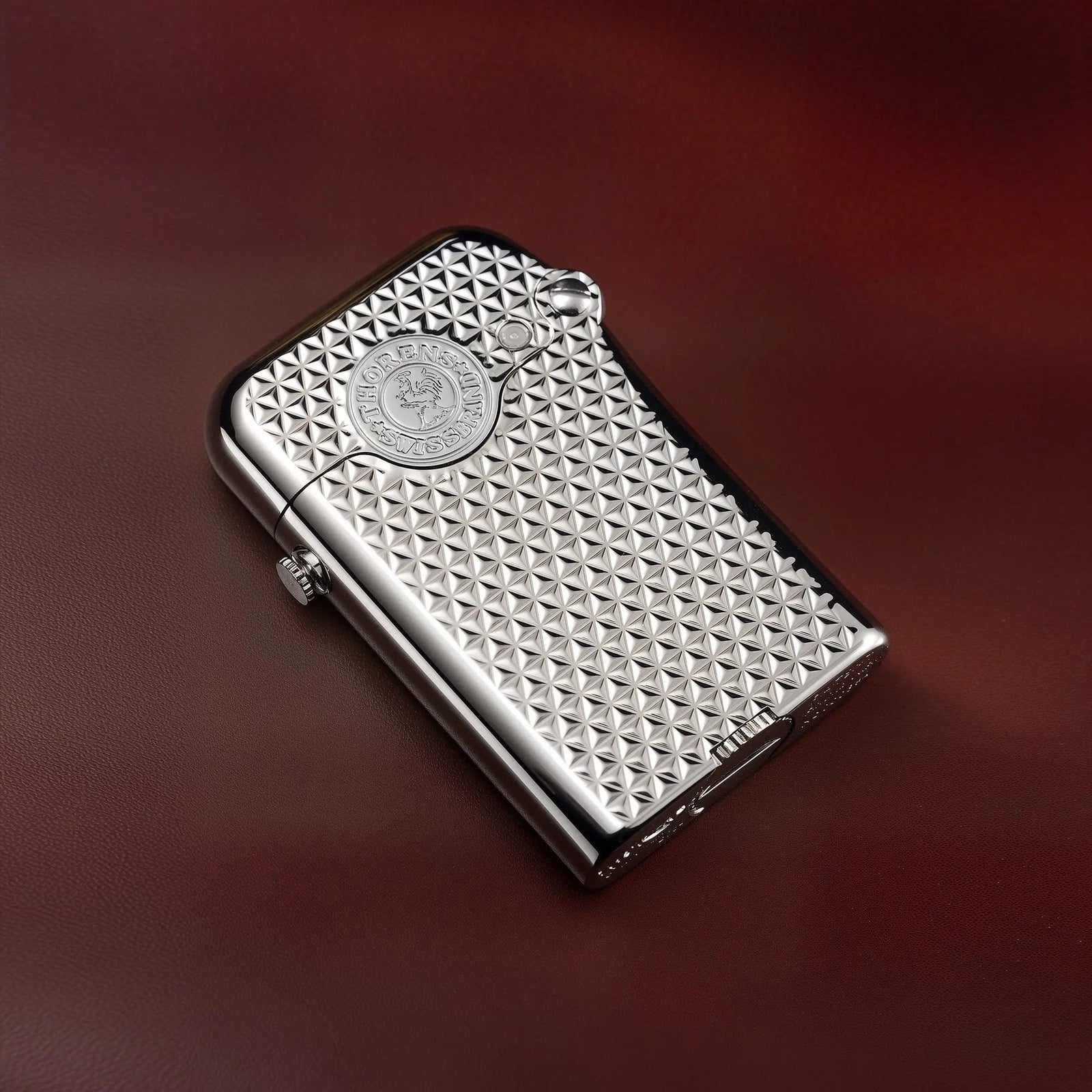 A Thorens Gen 3 Single Claw silver lighter featuring an intricate diamond grid texture and a central heritage emblem, resting on a smooth burgundy leather surface.
