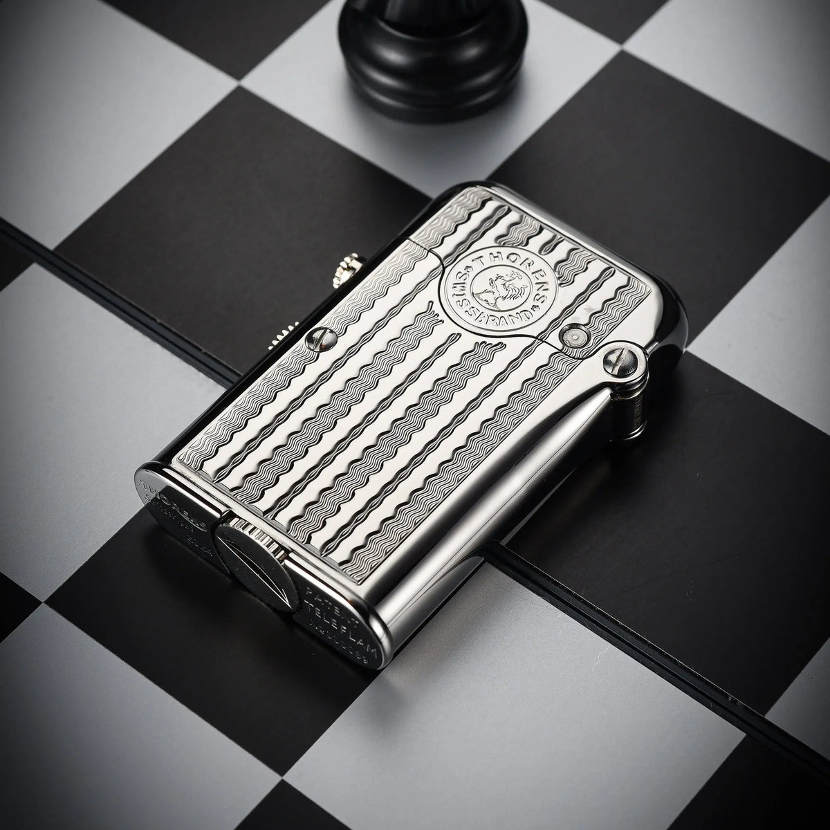 A silver Thorens Gen 7 TELEFLAM lighter featuring a sophisticated water ripple pattern and heritage emblem, resting on a black and white chessboard.