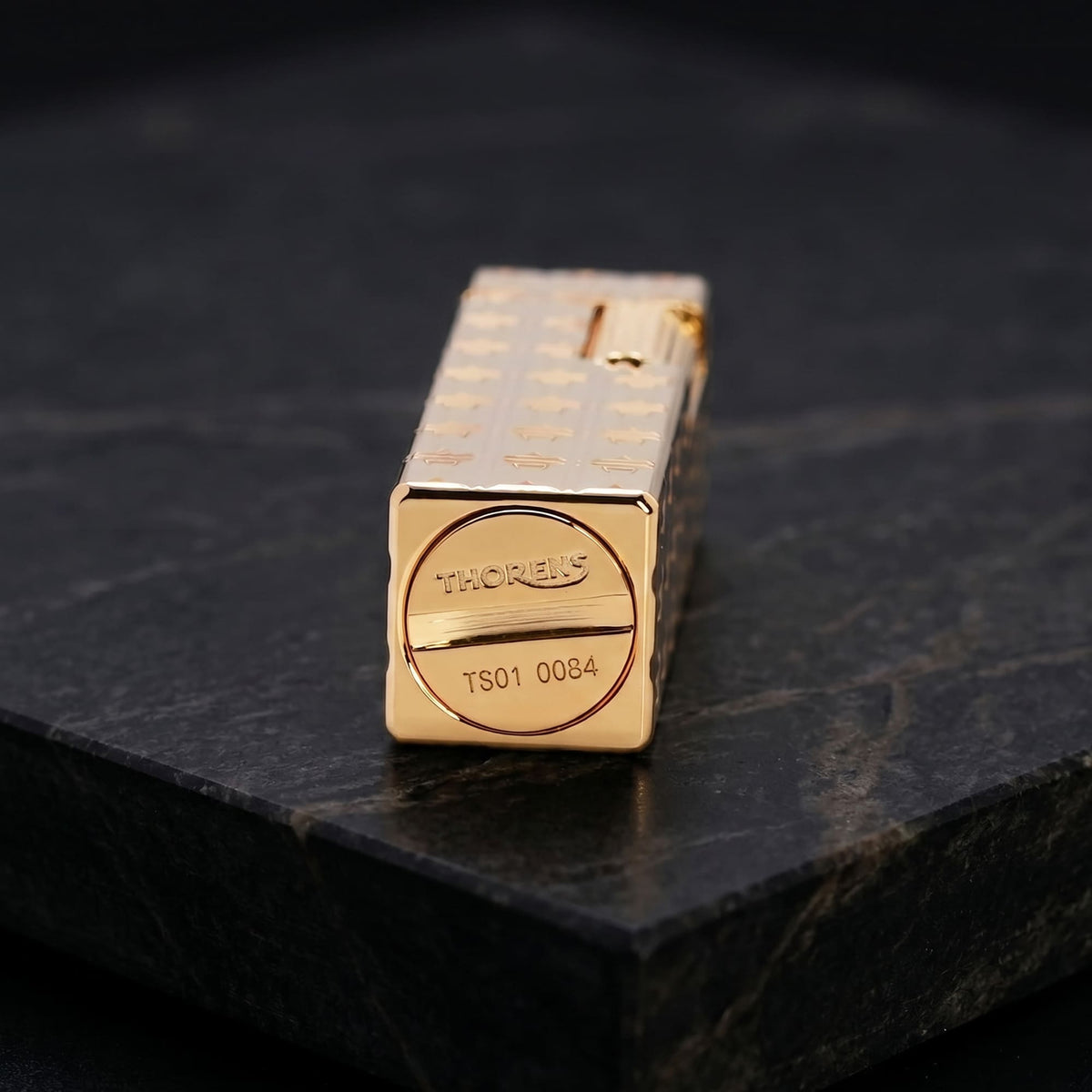 Close-up of the base of a Thorens Side Slip rectangular lighter with geometric block pattern and extended roller in gold finish, showing the engraved THORENS logo and serial number on a dark stone surface.