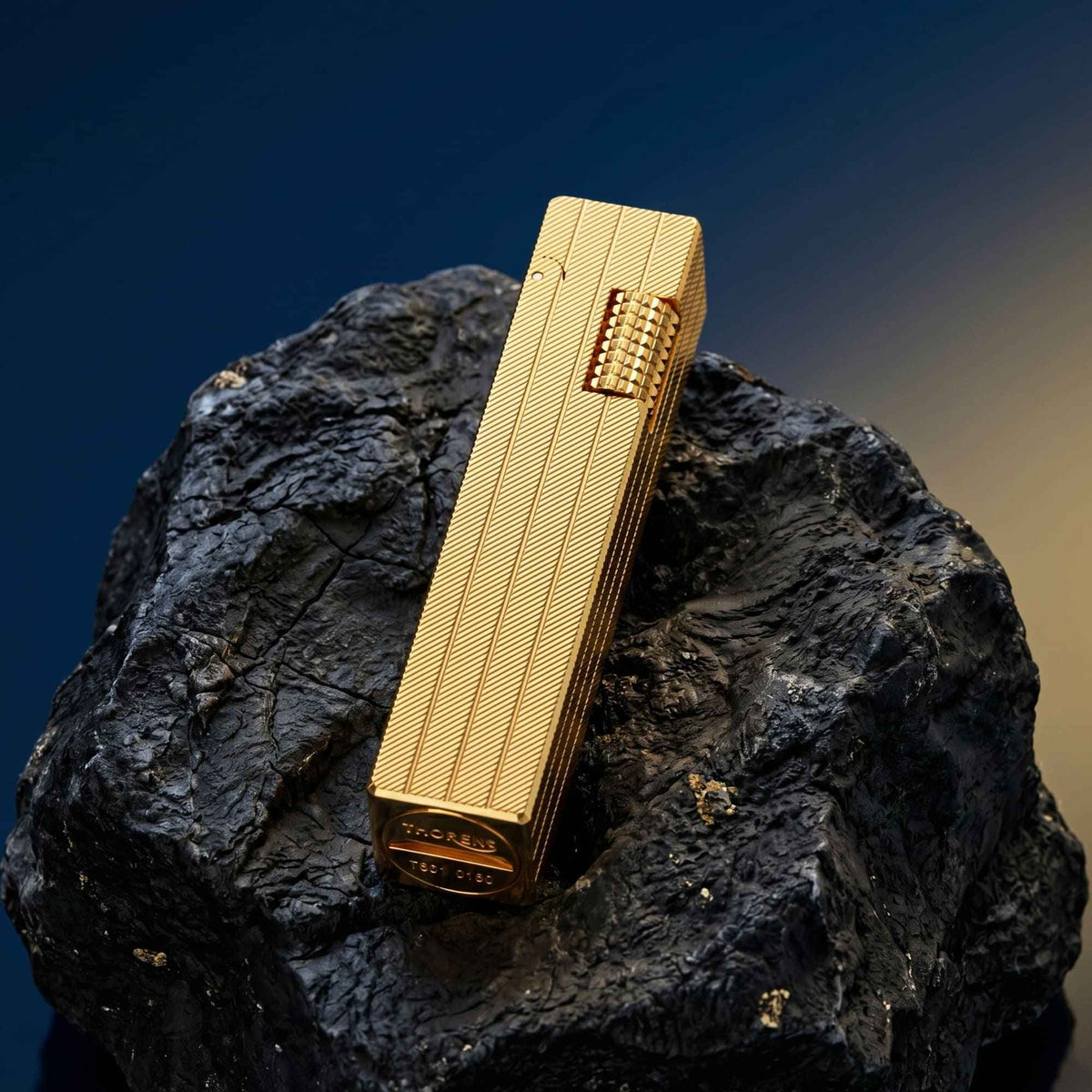 Thorens rectangular lighter with gold diagonal pattern shown with the lid down on dark stone, highlighting the surface pattern and side ignition wheel.