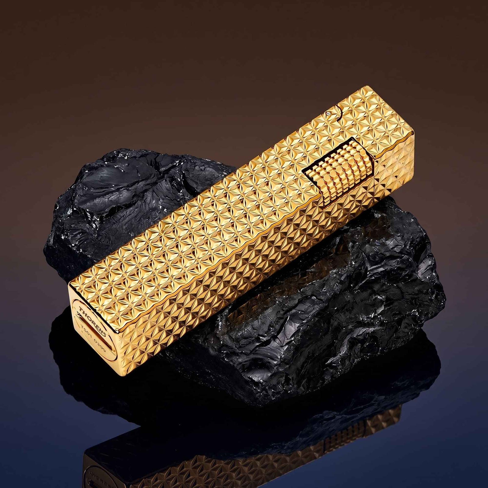 Thorens rectangular lighter with gold starburst pattern shown closed on dark stone, highlighting the raised surface pattern and side ignition wheel.
