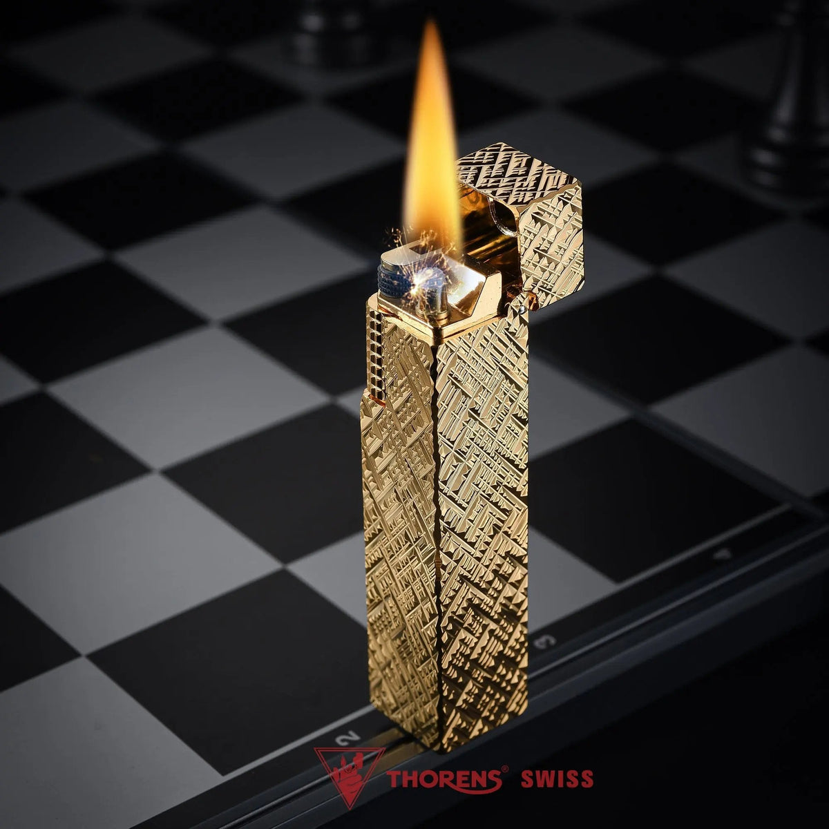 Lit gold carved rectangular Thorens side-slip lighter standing open on a black and white checkerboard surface