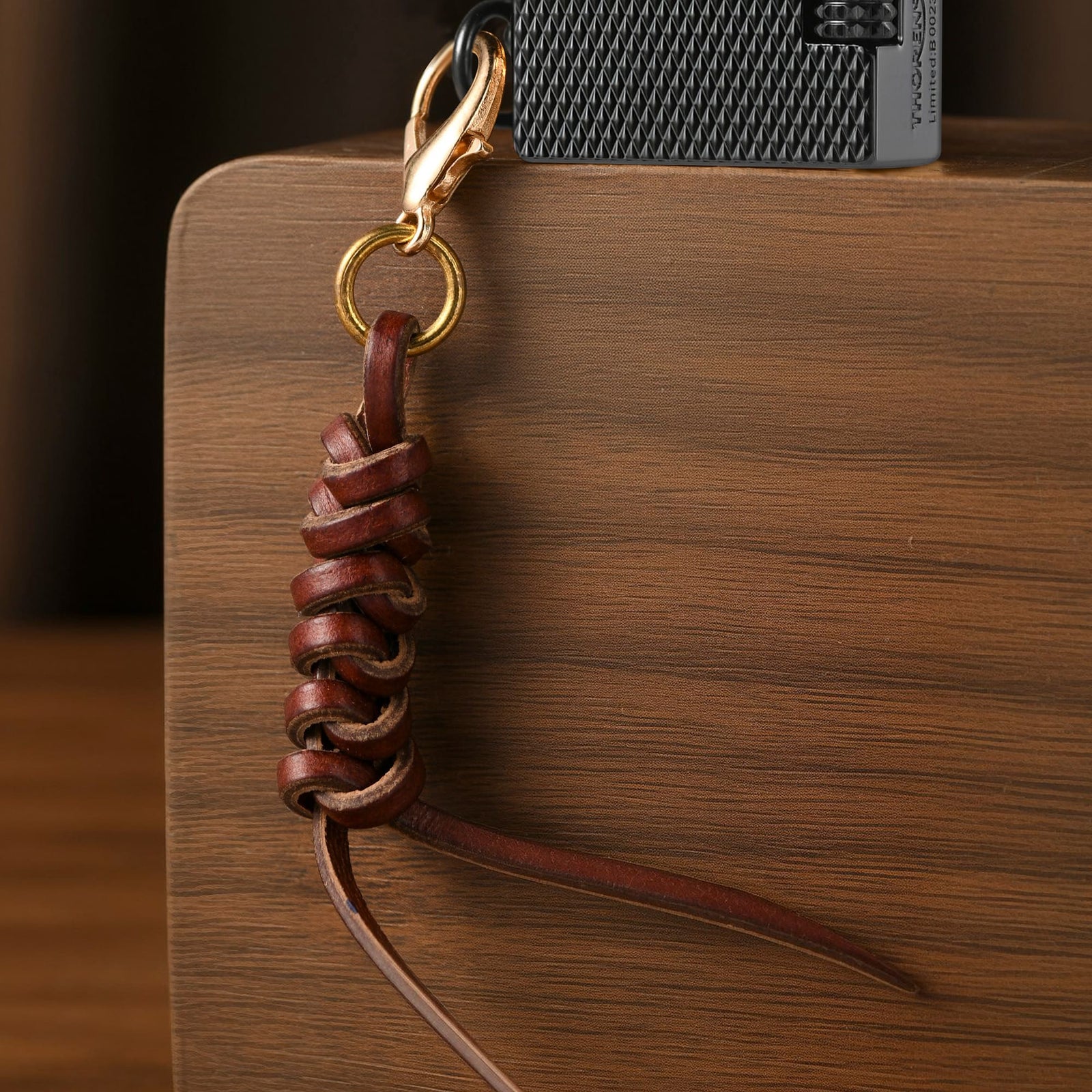 Handwoven cowhide leather charm with braided strap and metal clasp attached to the side of a lighter, showing natural leather texture on a wooden surface.