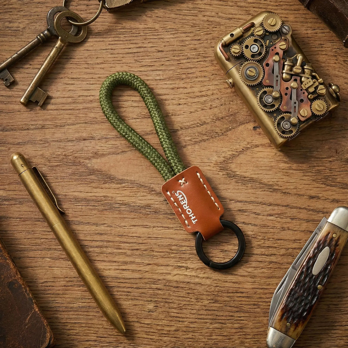 A Thorens green paracord keychain with a brown leather logo sleeve, surrounded by a brass pen, pocket knife, and gear-driven lighter on wood.