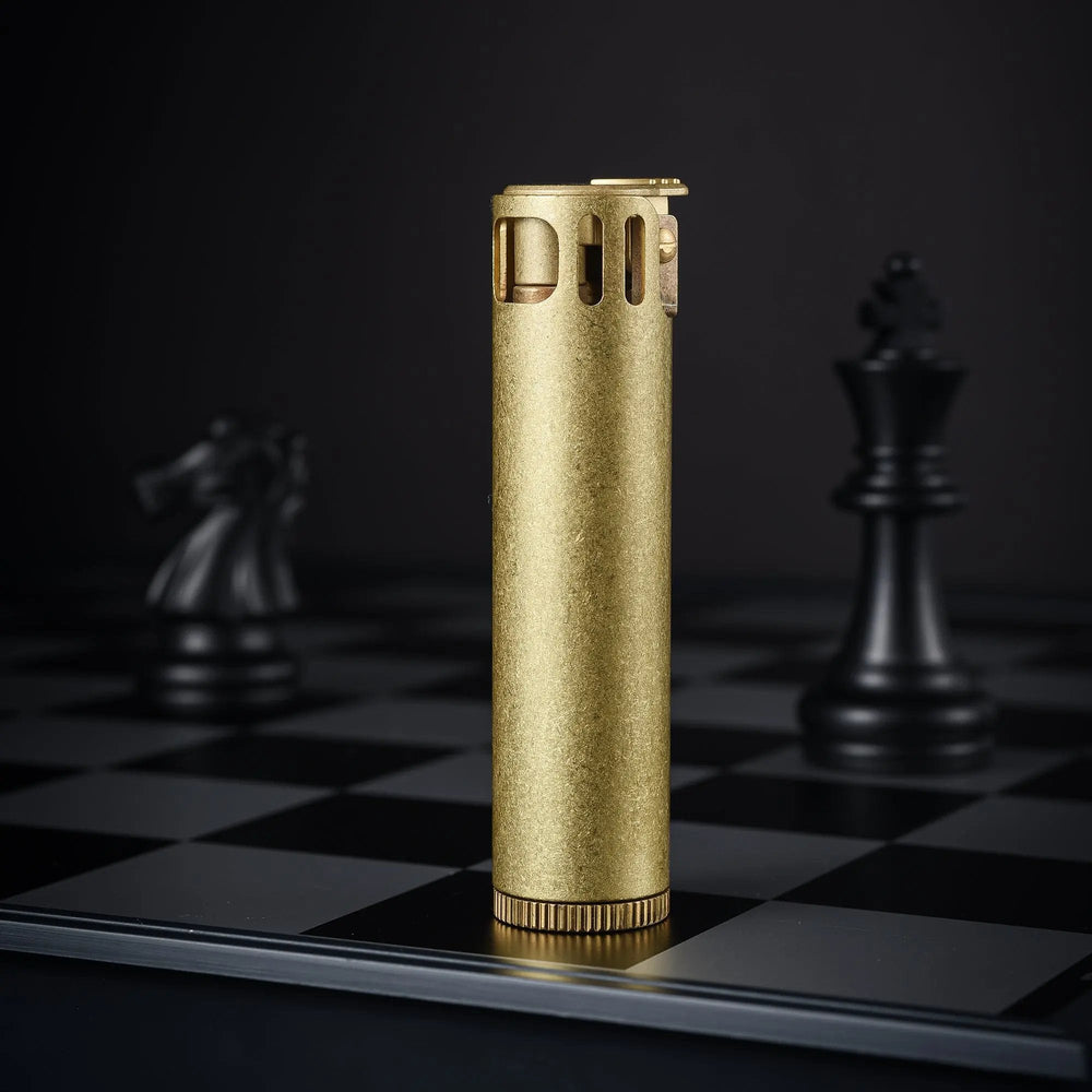 A vertical Ignis Tower lighter with a textured brass finish standing on a dark chessboard, showcasing its tall cylindrical design and slotted windguard.