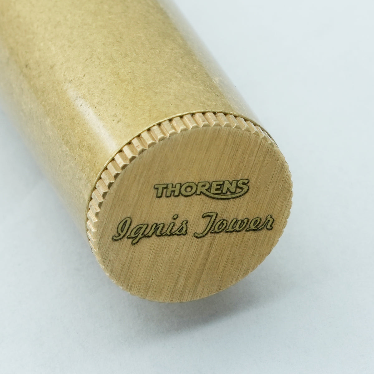 A vertical Ignis Tower lighter with a textured brass finish standing on a dark chessboard, showcasing its tall cylindrical design and slotted windguard.