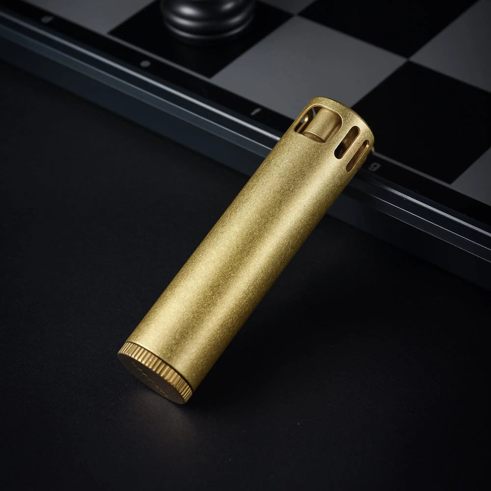 A brushed brass Ignis Tower lighter resting at an angle on a dark surface, with a black and white chessboard blurred in the background.