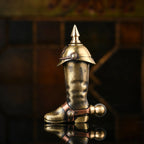 A side profile of the Knight's Boot permanent match lighter in aged brass, showing the textured boot surface, the spiked cap, and the spur-shaped flint wheel on a reflective dark surface.