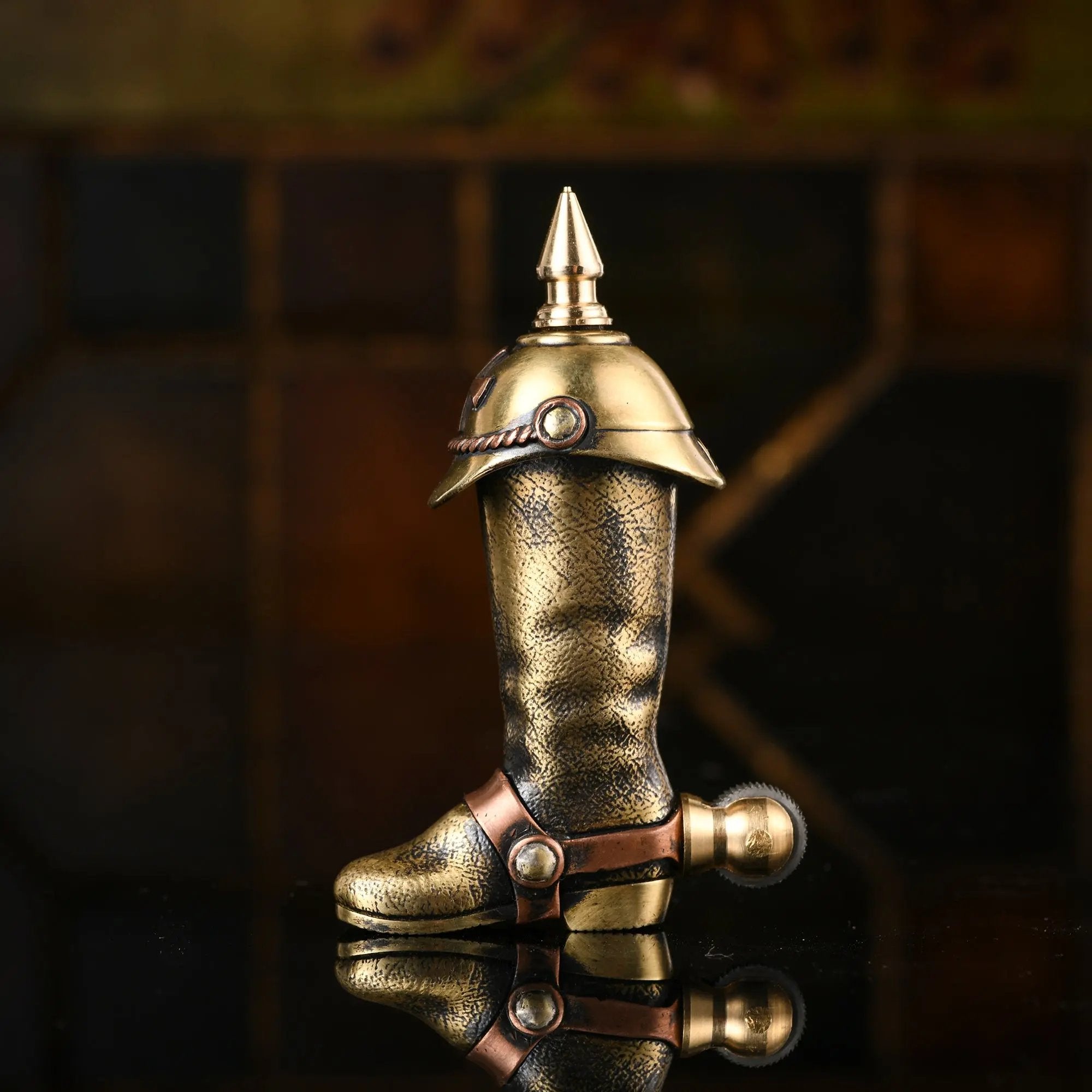 A side profile of the Knight's Boot permanent match lighter in aged brass, showing the textured boot surface, the spiked cap, and the spur-shaped flint wheel on a reflective dark surface.