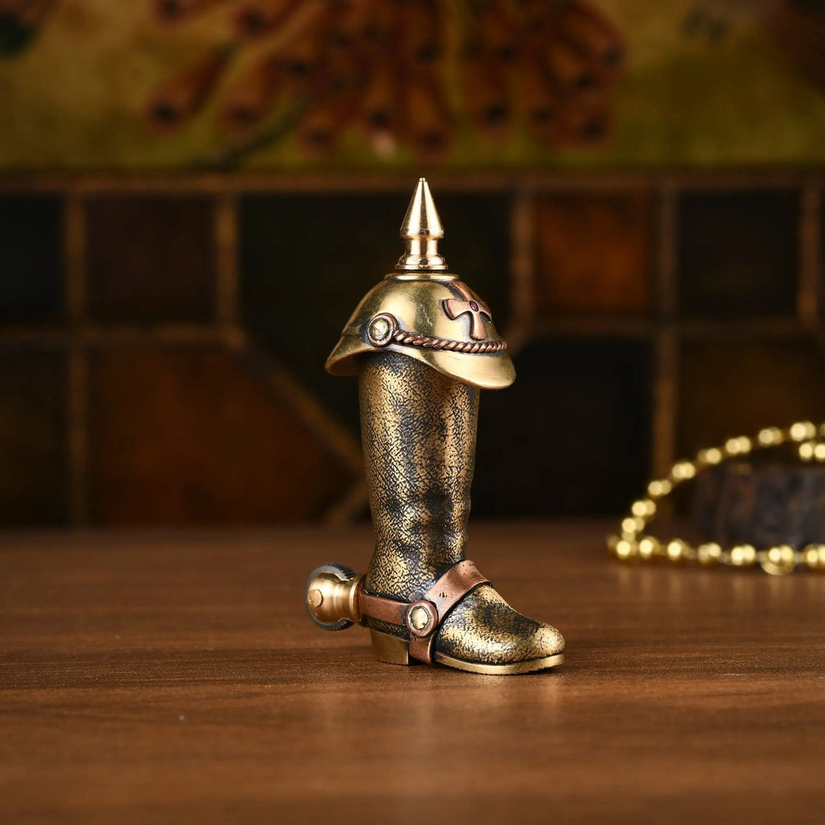 A full standing view of the Knight's Boot permanent match lighter in aged brass, featuring a pointed cap, cross emblem, and spur-shaped flint wheel, set on a rustic wooden surface.