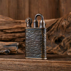 An intricately detailed, hand-carved Thorens Lockflame brass lighter featuring a rugged meteorite crater texture. The vintage mechanical ignition is visible, resting on a weathered wooden surface with wood art in the background.