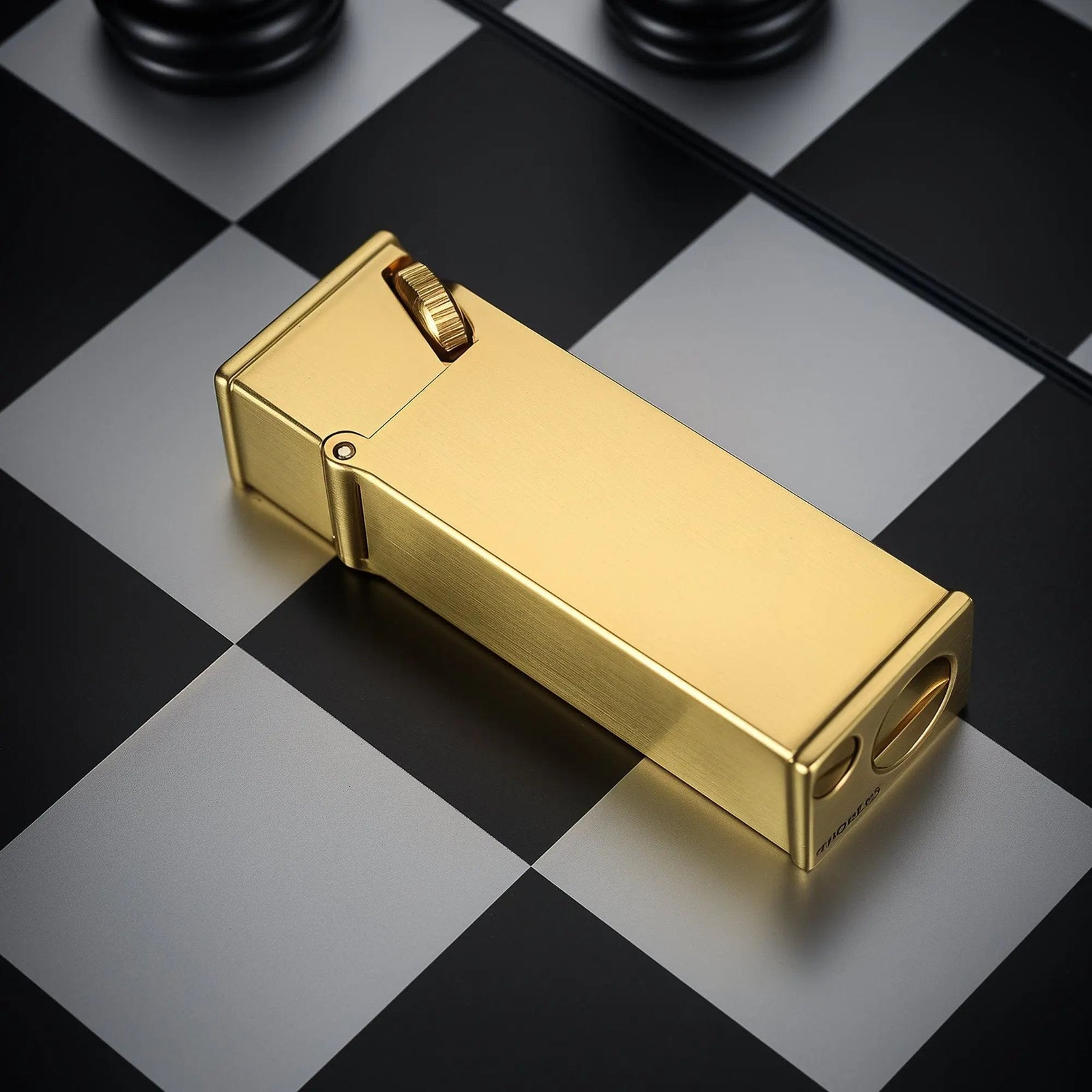 A brushed brass Long Fellow lighter resting horizontally on a black and white chessboard, showcasing its sleek rectangular body and inclined ignition wheel.