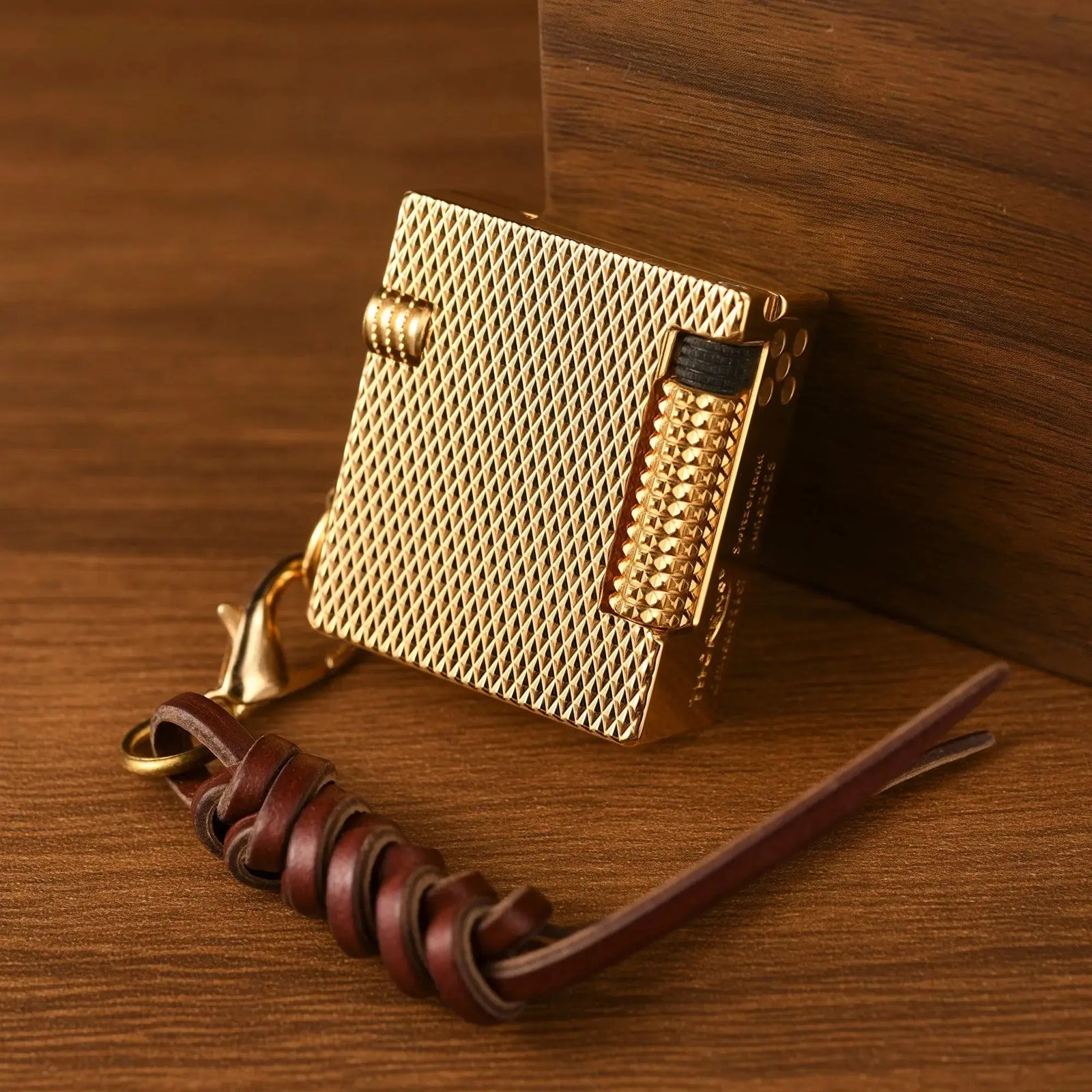 A compact Mini Cube Golden Mesh lighter with a knurled ignition roller and leather lanyard, resting on a warm wooden surface.