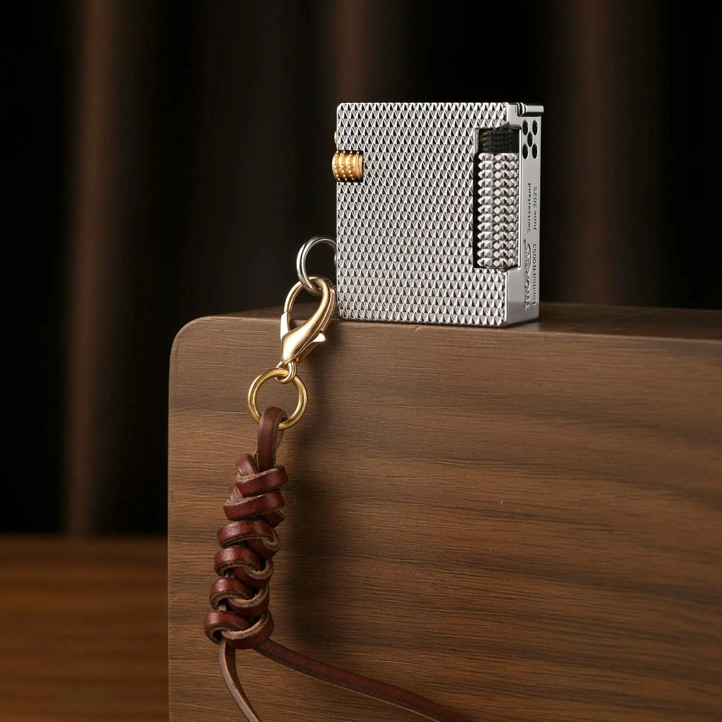 A Thorens Mini Cube lighter with a silver mesh texture and brass ignition wheel, standing upright on a wooden block with a leather lanyard.
