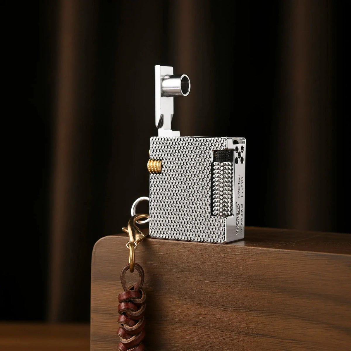 An open Thorens Mini Cube Silver Mesh lighter resting on a wooden surface, showing the lid mechanism and textured metal finish with a leather lanyard.