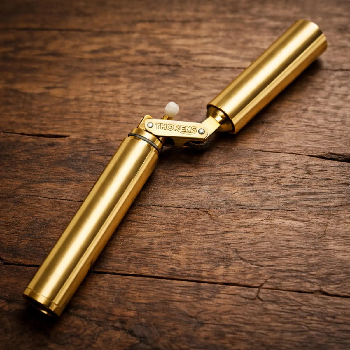 A Thorens Nunchaku lighter in a polished gold finish resting on a rustic wooden surface, showcasing its unique cylindrical "trench style" folding design and premium engraved brass hinge.