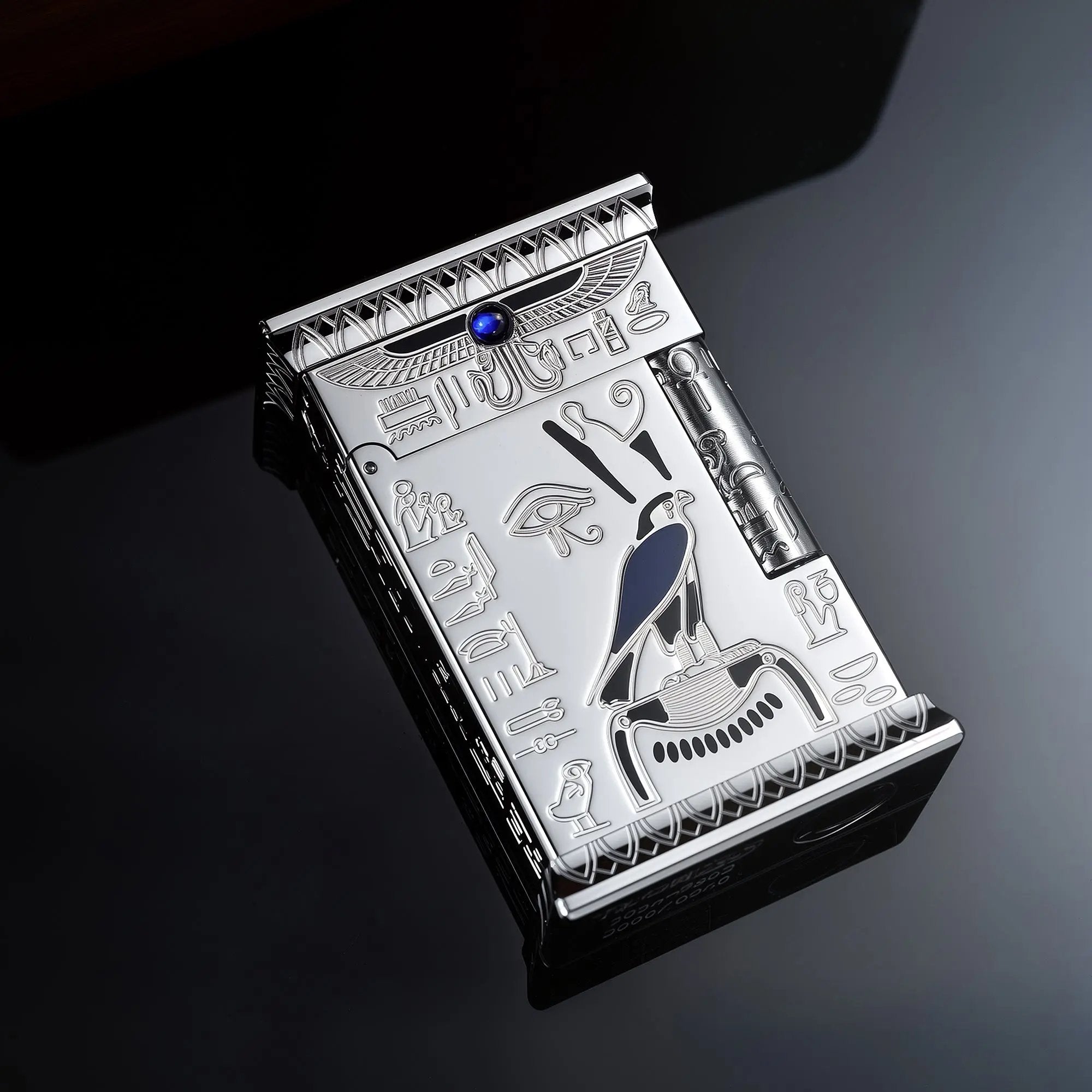 Polished silver Thorens lighter resting on a reflective surface, featuring detailed Egyptian falcon engravings, a blue gem, and hieroglyphs.