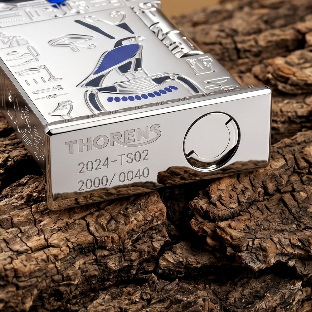 Polished silver Thorens lighter resting on a reflective surface, featuring detailed Egyptian falcon engravings, a blue gem, and hieroglyphs.
