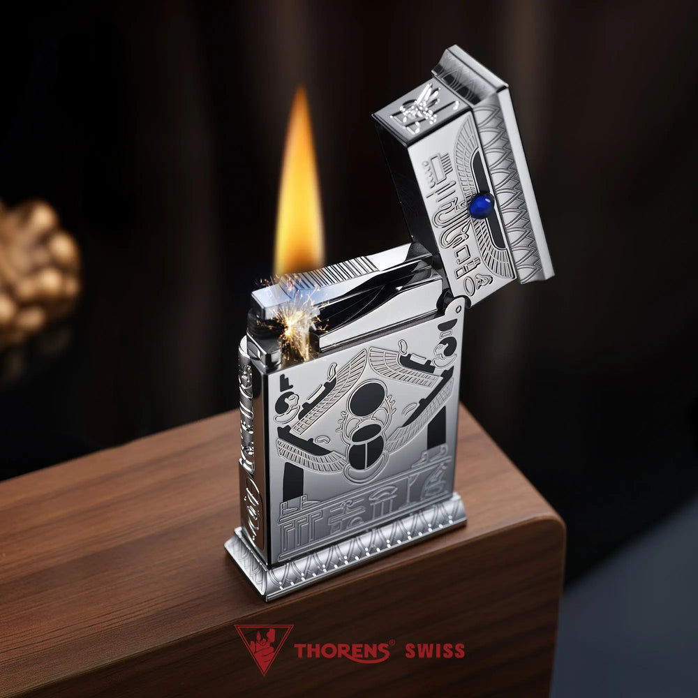 An open Pharaoh's Light Silver lighter ignited with a steady flame, showing a winged scarab engraving and blue gemstone on a metallic chrome body.