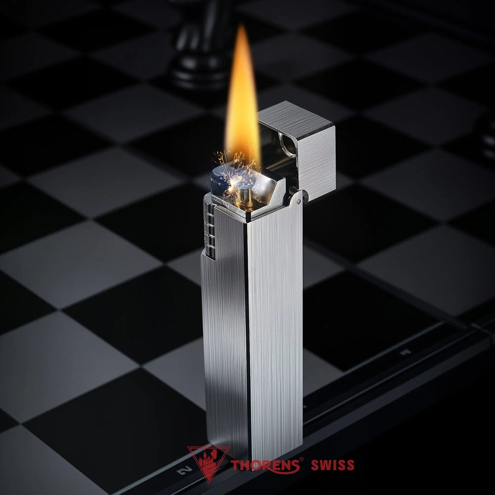 Glossy silver Thorens rectangular side-slip lighter standing upright with the lid open and flame lit, shown on a black and white checkered surface with the ignition wheel and wick clearly visible
