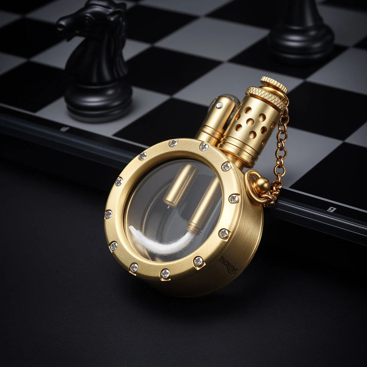 A round pocket watch lighter with a transparent fuel chamber and crystal-studded bezel, resting on a chessboard to showcase its mechanical brass craftsmanship.