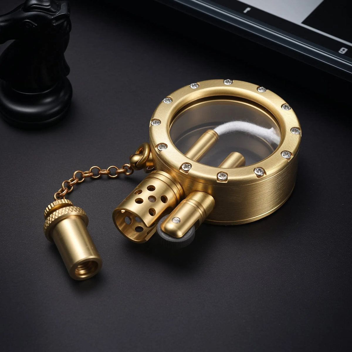 A brushed brass pocket watch lighter with an open cap, showing the flint wheel and perforated chimney next to a crystal-inlaid transparent fuel tank.