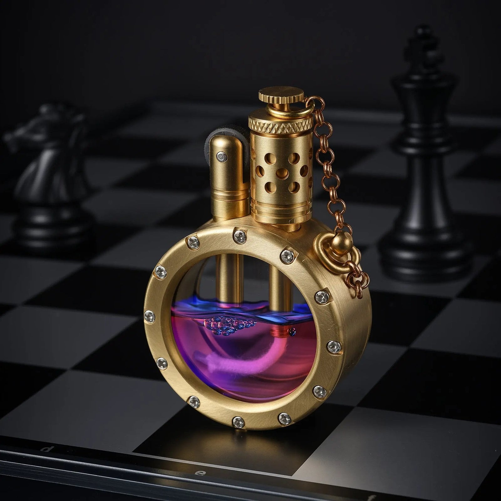 A brass finish round pocket watch lighter with a crystal-studded rim, filled with purple fuel, displayed on a black and white chessboard.