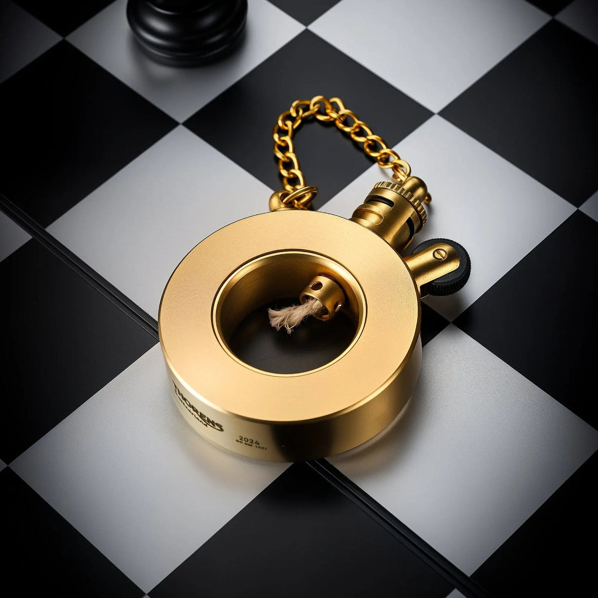 A Thorens Seahorse trench lighter with a unique circular brass body and gold-tone chain resting on a black and grey chessboard.
