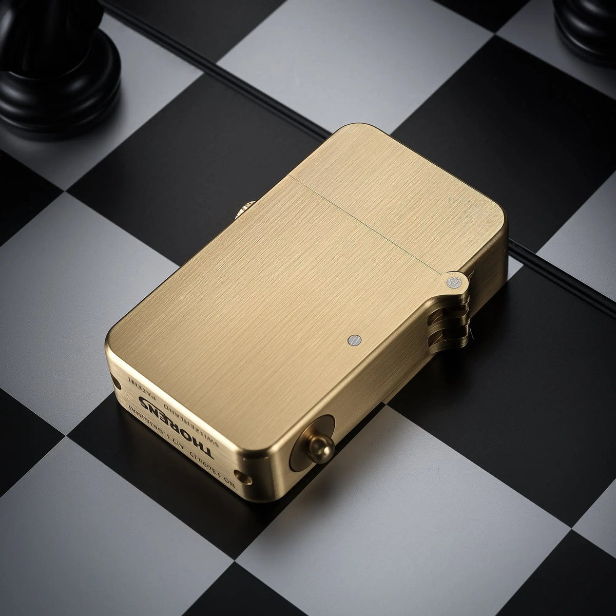 A Thorens semi-automatic ignition lighter with a brushed glossy brass finish rests on a black and grey chessboard, showcasing its heritage mechanical design.