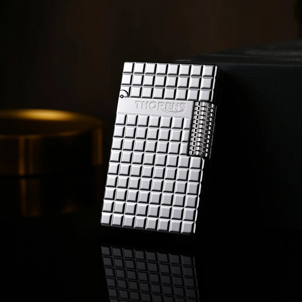 A silver Thorens side slip lighter leaning against a black box, showcasing the reflective argent block pattern and precision side-roller ignition.