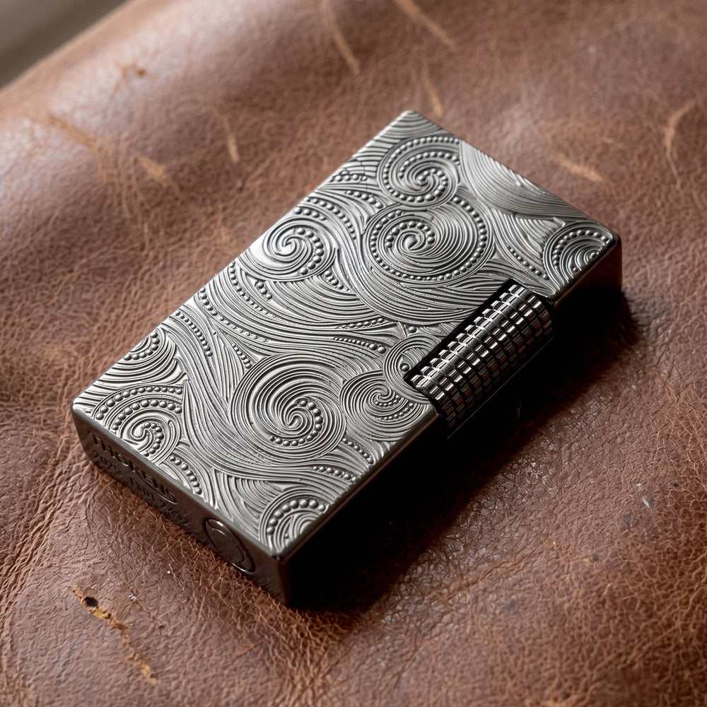A close-up perspective of the Thorens Side Slip lighter in black with a Vortex engraved pattern, resting on a dark stone surface. The shot highlights the precision-engraved "THORENS" logo and model serial number on the base, emphasizing its mechanical precision and collectible quality.