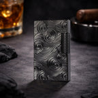 A professional lifestyle shot of the black Thorens Side Slip lighter with a deep Vortex engraved texture, standing upright on a dark slate surface. The background features a glass of whiskey and a cigar humidor, emphasizing its heritage appeal and premium EDC collectible status.