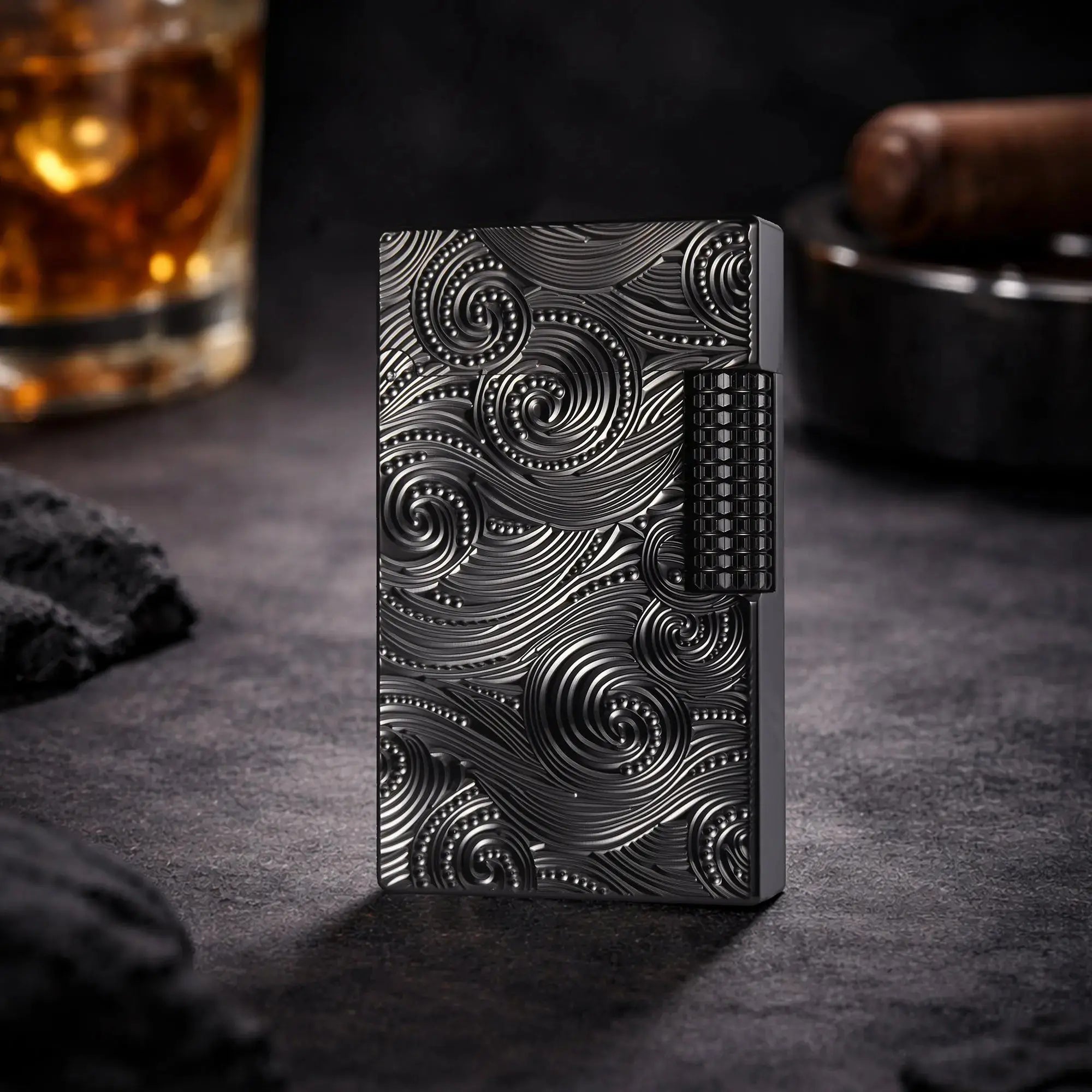 A professional lifestyle shot of the black Thorens Side Slip lighter with a deep Vortex engraved texture, standing upright on a dark slate surface. The background features a glass of whiskey and a cigar humidor, emphasizing its heritage appeal and premium EDC collectible status.