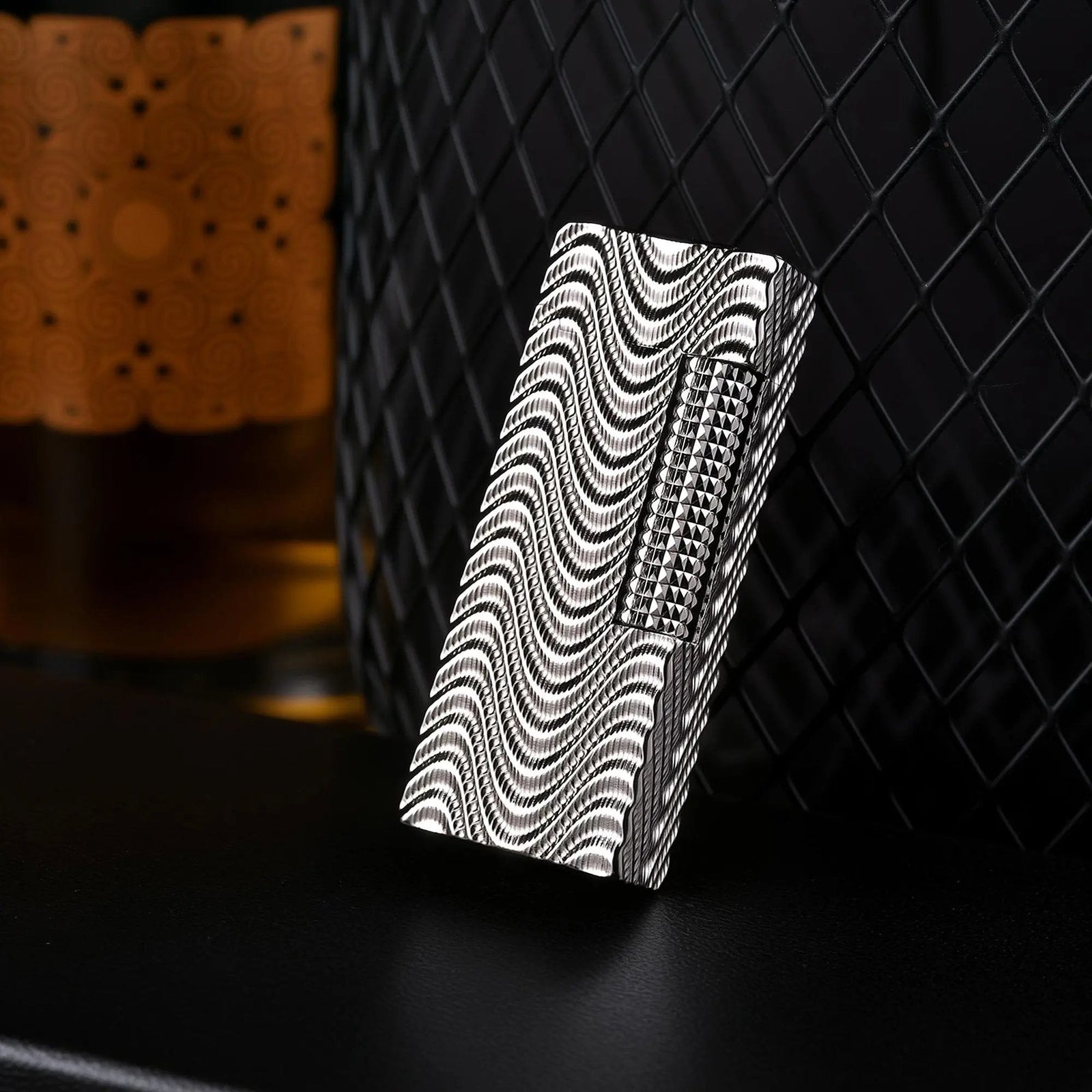 An Argent Mirage narrow lighter angled diagonally on a black surface, showcasing its high-density silver wave texture against a background of black wire mesh and warm ambient lighting.