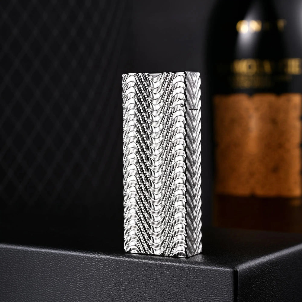 A vertical portrait of the Argent Mirage narrow lighter, featuring a dense, silver wave-textured pattern that creates a shimmering optical effect, standing on a black surface with a blurred wine bottle in the background.