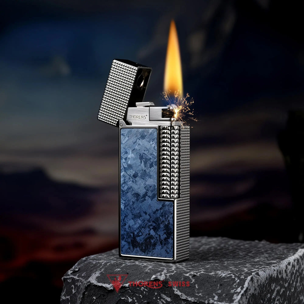 An ignited Blue Frost Ti narrow lighter with a crystallized blue inlay and silver hobnail texture, featuring a bright flame and golden sparks against a scenic mountain backdrop.