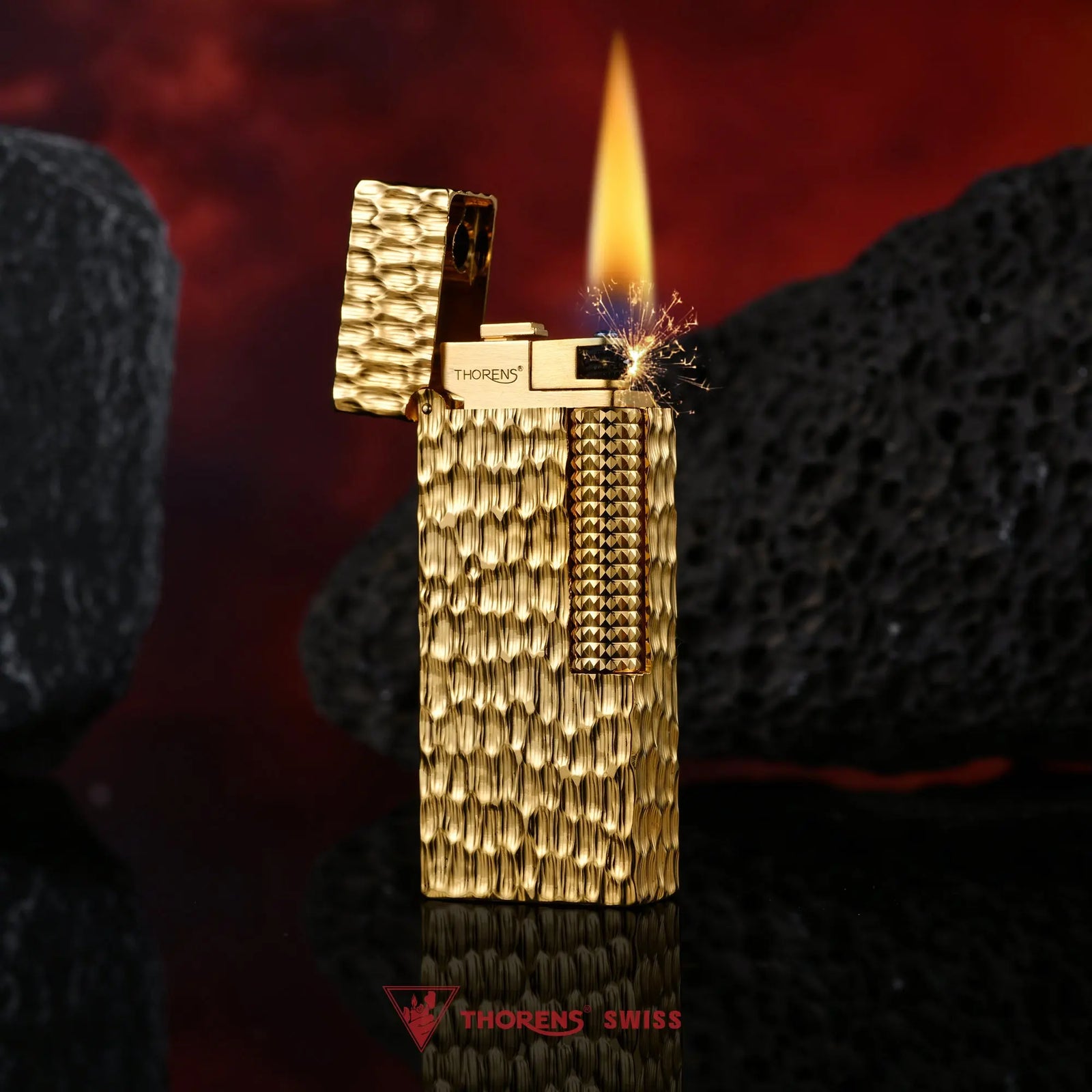 An ignited Golden Crater narrow lighter with an open lid, showing a warm flame and sparks against a hammered gold surface and dark volcanic stones.