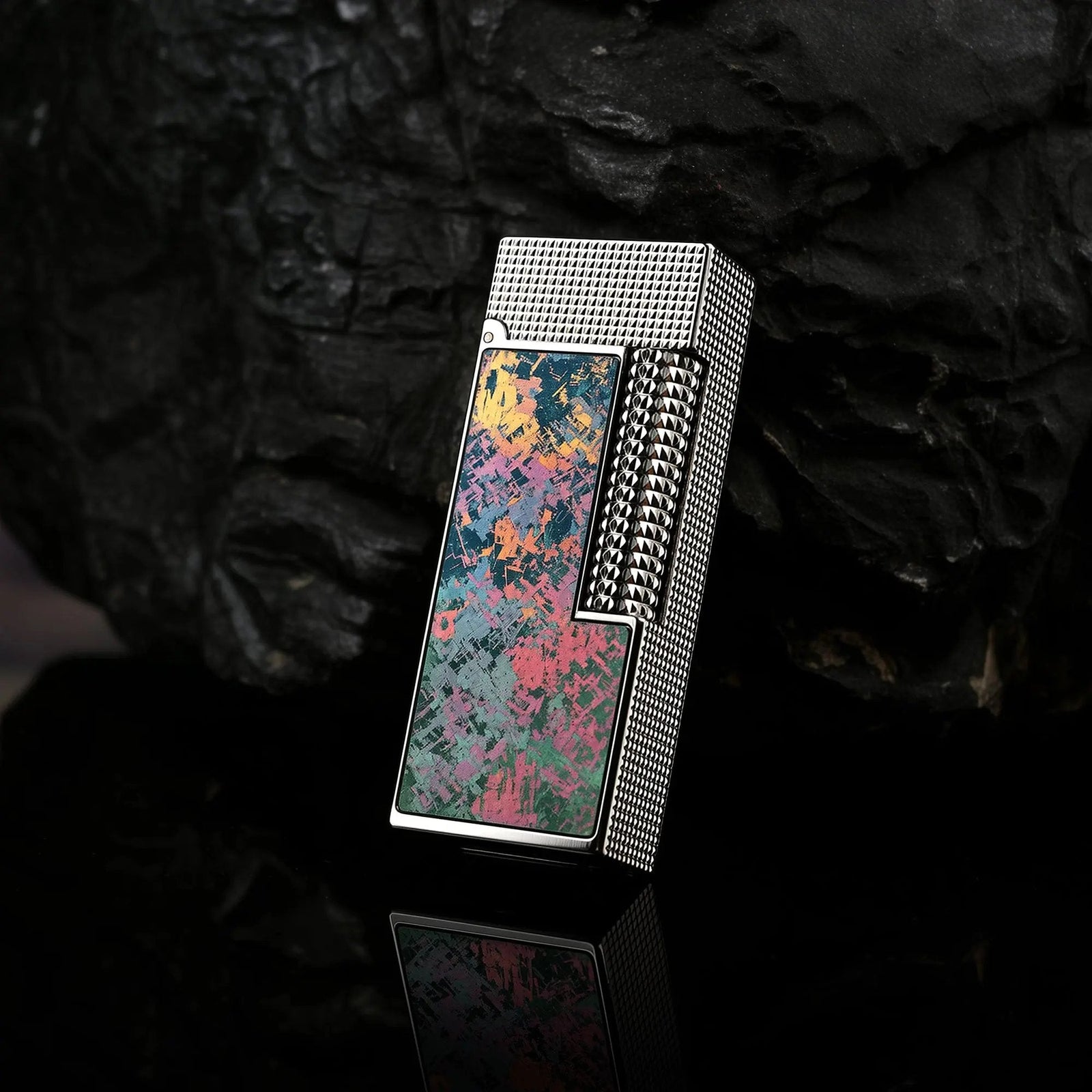 A striking profile of the Iridescent Ti narrow lighter resting against a dark, textured coal background, highlighting the vivid pink, teal, and gold splashes on its titanium faceplate.