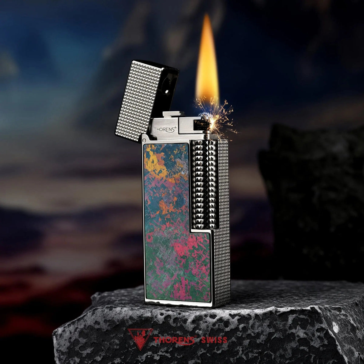 An open Iridescent Ti narrow lighter in mid-ignition, throwing sparks and a warm flame that contrasts with its colorful pink and green titanium inlay and silver hobnail body.