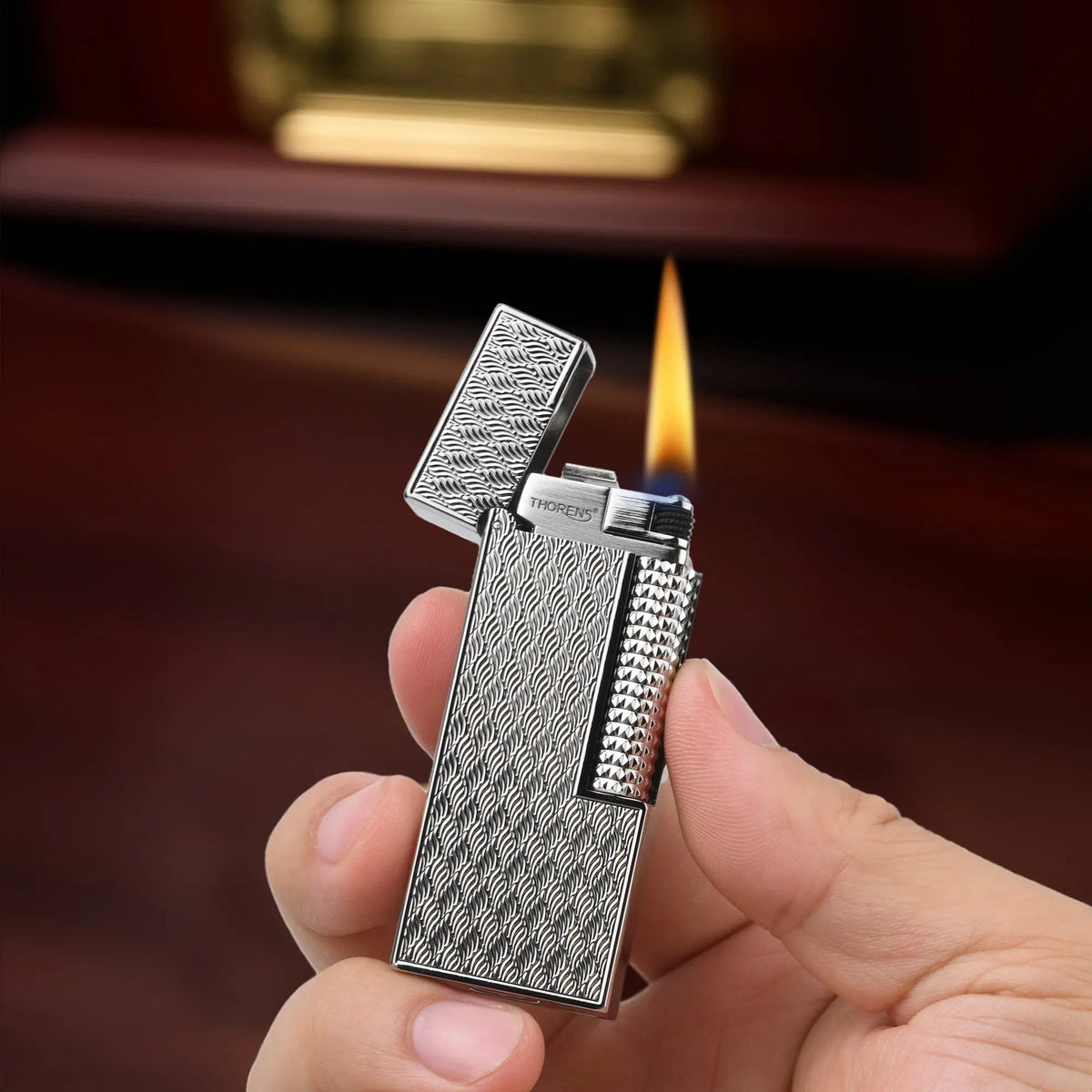 A person holding the ignited Ripple Shadow narrow lighter, showcasing its silver-toned wavy texture and vertical flame against a warm, blurred wooden background.