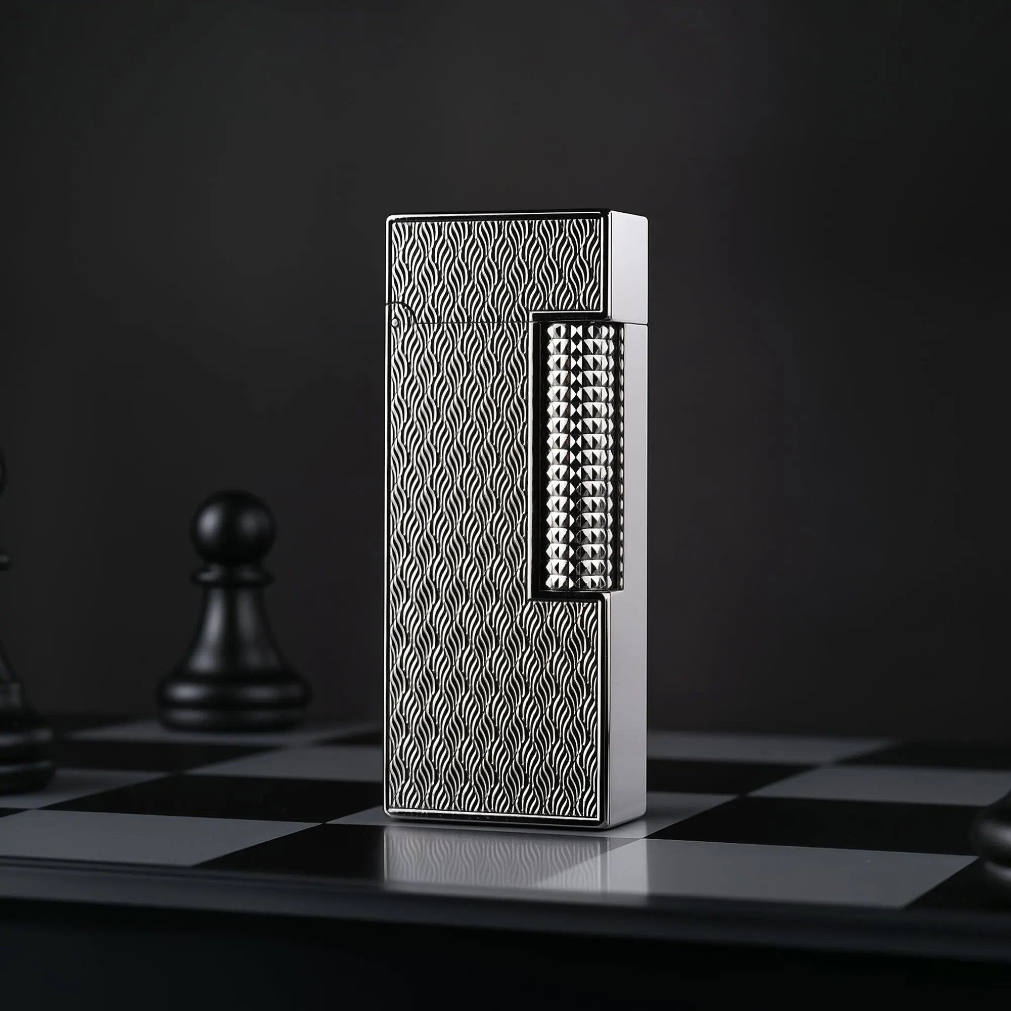 A vertical product shot of the Ripple Shadow narrow lighter standing on a chessboard. The silver-toned wavy texture of the lighter is in sharp focus, contrasted against a dark background with a blurred black chess pawn.