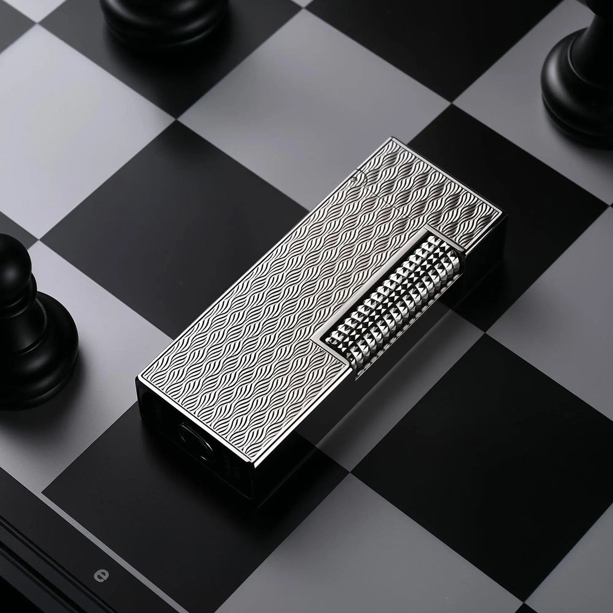 A Ripple Shadow narrow lighter lying diagonally on a black and grey chessboard, showcasing its silver-toned wavy "ripple" texture and polished hobnail ignition roller.